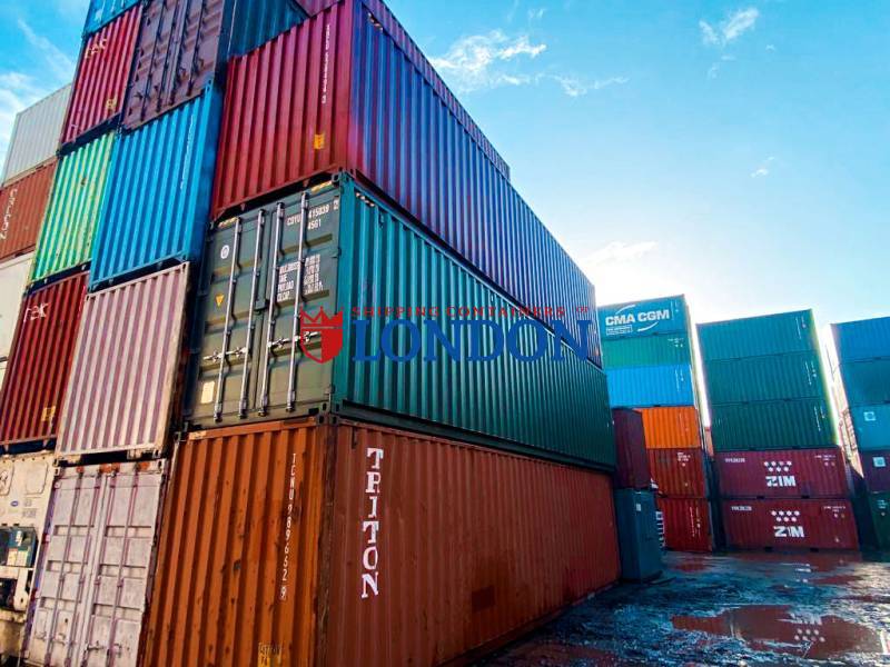A stack of colorful shipping containers against a blue sky, reflecting light on a wet surface.