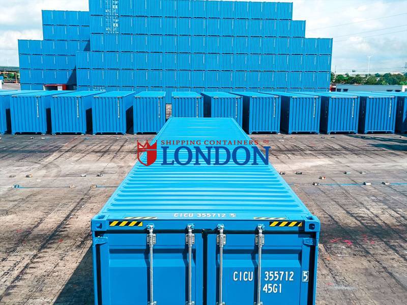 A stack of blue shipping containers arranged in a storage area under a cloudy sky.